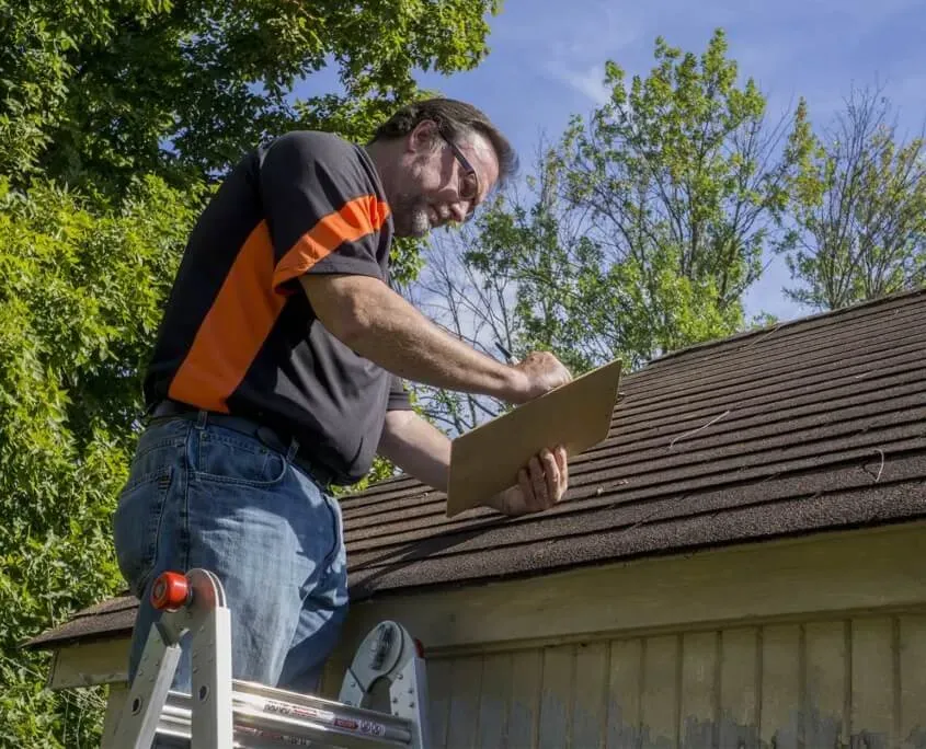 Man on ladder looking at rooftop for hail damage and recording his findings on a notepad